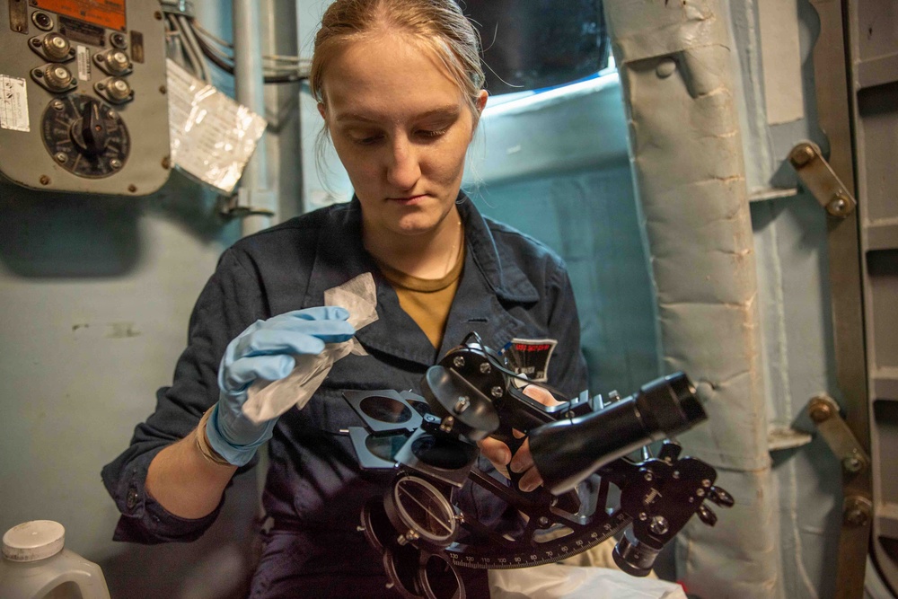 A young woman in a dark blue naval uniform is carefully cleaning or maintaining a marine sextant while wearing blue latex gloves. She is working in what appears to be a shipboard interior environment, surrounded by naval equipment and bulkhead fixtures. The activity suggests routine maintenance of navigational instruments aboard a military vessel.