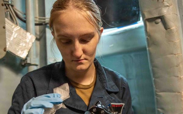 A young woman in a dark blue naval uniform is carefully cleaning or maintaining a marine sextant while wearing blue latex gloves. She is working in what appears to be a shipboard interior environment, surrounded by naval equipment and bulkhead fixtures. The activity suggests routine maintenance of navigational instruments aboard a military vessel.