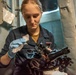 A young woman in a dark blue naval uniform is carefully cleaning or maintaining a marine sextant while wearing blue latex gloves. She is working in what appears to be a shipboard interior environment, surrounded by naval equipment and bulkhead fixtures. The activity suggests routine maintenance of navigational instruments aboard a military vessel.