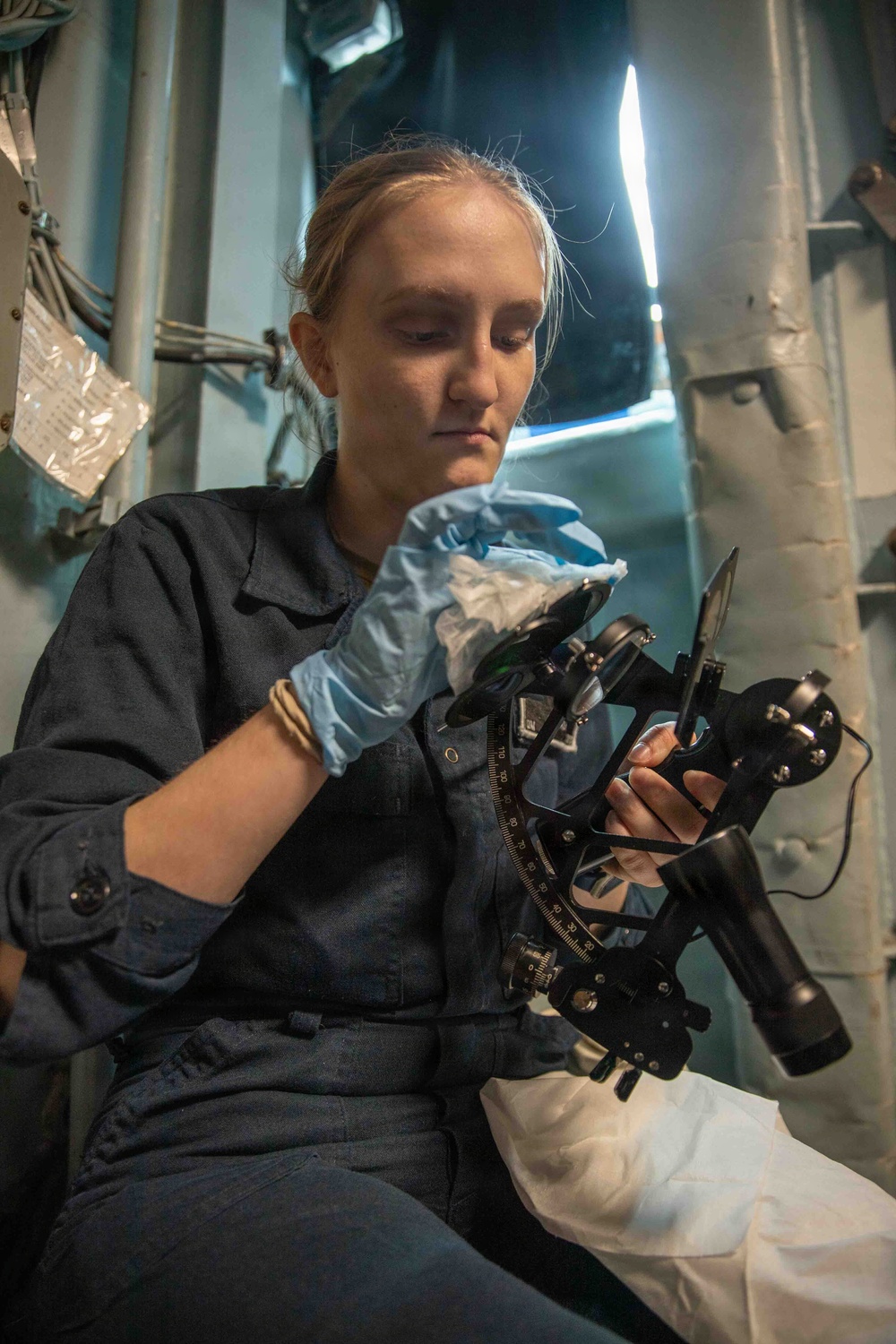 A young woman in a dark navy uniform is carefully cleaning or maintaining a nautical sextant using a cloth while wearing blue latex gloves. She appears to be seated in a confined shipboard space, suggesting a military or maritime vessel environment. The image captures a maintenance or inspection task being performed on precision navigational equipment.