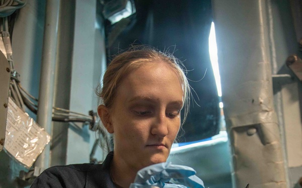 A young woman in a dark navy uniform is carefully cleaning or maintaining a nautical sextant using a cloth while wearing blue latex gloves. She appears to be seated in a confined shipboard space, suggesting a military or maritime vessel environment. The image captures a maintenance or inspection task being performed on precision navigational equipment.