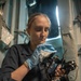 A young woman in a dark navy uniform is carefully cleaning or maintaining a nautical sextant using a cloth while wearing blue latex gloves. She appears to be seated in a confined shipboard space, suggesting a military or maritime vessel environment. The image captures a maintenance or inspection task being performed on precision navigational equipment.
