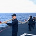 Naval personnel in dark blue uniforms are handling a large mooring or tow rope on the deck of a military vessel at sea. At least three crew members are visible working together to manage the thick braided rope. The ship's superstructure is visible on the right side, and the open ocean can be seen in the background.