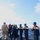 A group of naval personnel aboard a warship are collectively carrying a large rope or hawser line across the deck. The crew members are wearing standard navy working uniforms and colored safety helmets, suggesting an organized deck evolution or replenishment-at-sea (RAS) operation. The vessel is underway on open ocean under partly cloudy skies.