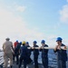 A group of naval personnel aboard a warship are collectively carrying a large rope or hawser line across the deck. The crew members are wearing standard navy working uniforms and colored safety helmets, suggesting an organized deck evolution or replenishment-at-sea (RAS) operation. The vessel is underway on open ocean under partly cloudy skies.