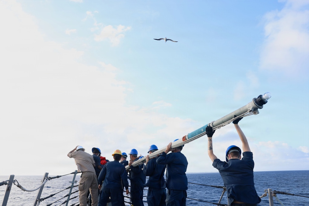 A group of naval personnel aboard a ship at sea are collectively handling and lifting a large cylindrical object, likely a torpedo or missile, on the deck. The crew members are wearing standard naval working uniforms and colored hard hats indicating different roles or departments. A seabird is visible flying overhead in a partly cloudy sky.