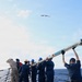 A group of naval personnel aboard a ship at sea are collectively handling and lifting a large cylindrical object, likely a torpedo or missile, on the deck. The crew members are wearing standard naval working uniforms and colored hard hats indicating different roles or departments. A seabird is visible flying overhead in a partly cloudy sky.