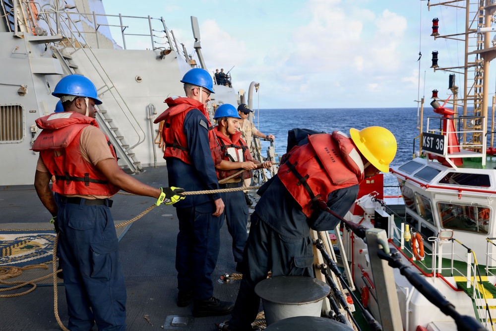 Naval personnel are conducting a replenishment-at-sea or vessel transfer operation between two ships on open ocean. Four crew members wearing red life vests and blue or yellow hard hats are handling lines and ropes on the deck. A smaller vessel marked 'KST 36' is positioned alongside the larger gray naval ship.