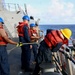 Naval personnel are conducting a replenishment-at-sea or vessel transfer operation between two ships on open ocean. Four crew members wearing red life vests and blue or yellow hard hats are handling lines and ropes on the deck. A smaller vessel marked 'KST 36' is positioned alongside the larger gray naval ship.