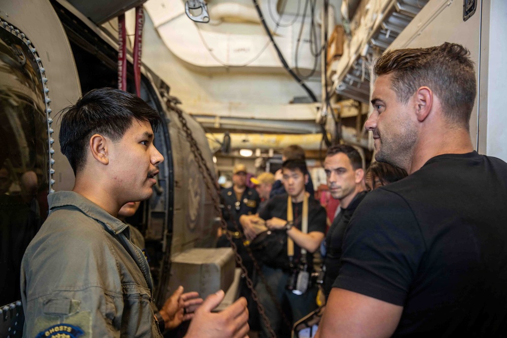 A group of people are gathered in a narrow industrial corridor, likely aboard a naval vessel or ship. Two men in the foreground appear to be engaged in conversation, with one wearing a military-style flight suit patch labeled 'GHOSTS'. Several other individuals are visible in the background observing or listening.