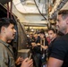 A group of people are gathered in a narrow industrial corridor, likely aboard a naval vessel or ship. Two men in the foreground appear to be engaged in conversation, with one wearing a military-style flight suit patch labeled 'GHOSTS'. Several other individuals are visible in the background observing or listening.