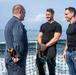Three men are engaged in a friendly conversation on the deck of a naval vessel at sea. One man on the left is wearing a navy blue military coverall uniform, while the other two are dressed in black civilian t-shirts. The ocean is visible in the background, and a safety net lines the ship's railing.