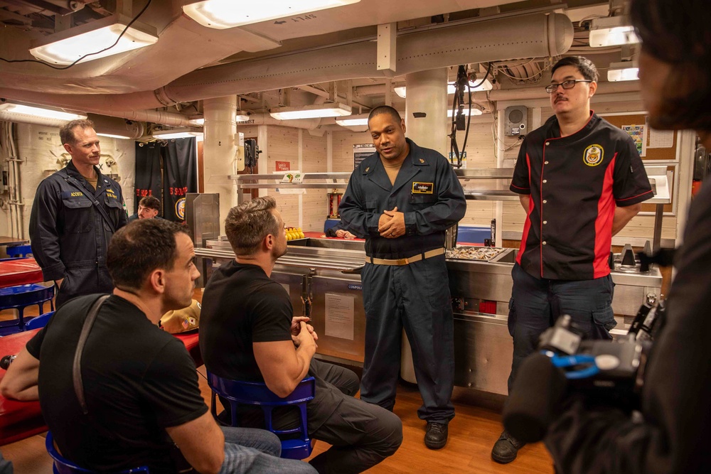 A group of people are gathered in what appears to be a military ship's galley or mess hall. A person in a blue naval coverall uniform is speaking to seated civilians, while another individual in a black chef's uniform with a military insignia patch stands nearby. The setting suggests a tour, briefing, or media visit aboard a U.S. Navy vessel.