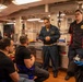 A group of people are gathered in what appears to be a military ship's galley or mess hall. A person in a blue naval coverall uniform is speaking to seated civilians, while another individual in a black chef's uniform with a military insignia patch stands nearby. The setting suggests a tour, briefing, or media visit aboard a U.S. Navy vessel.