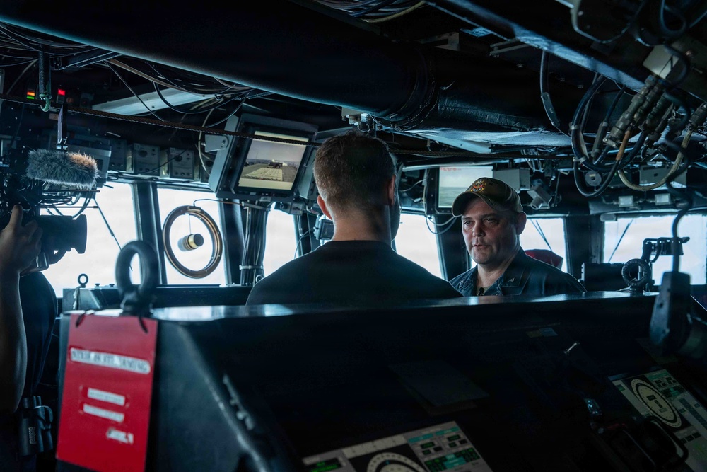 The image shows the bridge or combat information center of a naval vessel, where two individuals appear to be engaged in a conversation or interview. A camera crew with a boom microphone is visible on the left side, suggesting a media or documentary recording is taking place. The environment is filled with navigation and control equipment typical of a military ship's bridge.