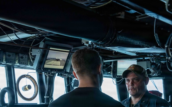 The image shows the bridge or combat information center of a naval vessel, where two individuals appear to be engaged in a conversation or interview. A camera crew with a boom microphone is visible on the left side, suggesting a media or documentary recording is taking place. The environment is filled with navigation and control equipment typical of a military ship's bridge.