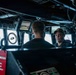 The image shows the bridge or combat information center of a naval vessel, where two individuals appear to be engaged in a conversation or interview. A camera crew with a boom microphone is visible on the left side, suggesting a media or documentary recording is taking place. The environment is filled with navigation and control equipment typical of a military ship's bridge.