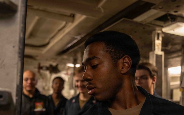 A young sailor in dark Navy working uniform is handling what appears to be an artillery shell or projectile aboard a naval vessel. Several other personnel are visible in the background observing or participating in the activity. The setting appears to be an interior compartment or magazine area of a warship.