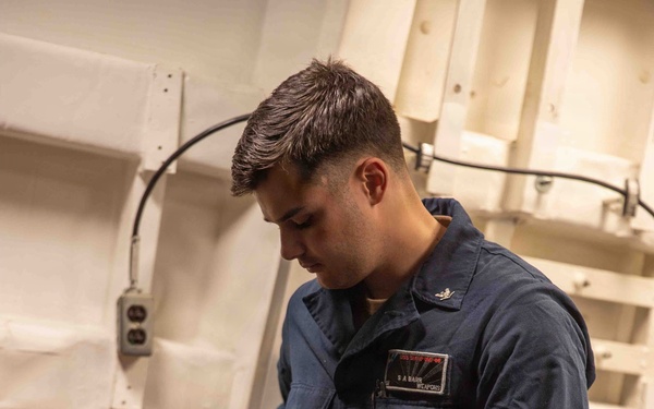 A U.S. Navy sailor in blue coveralls (NWU Type III or similar working uniform) is handling a firearm, likely during an armory or weapons handling procedure. The individual wears a name tape reading 'S A BARR' and a 'WEAPONS' department patch, along with a duty belt. The setting appears to be an interior shipboard compartment or armory space.
