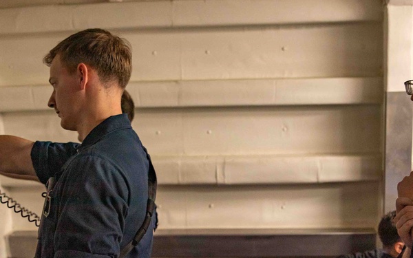 Two individuals in dark blue military-style uniforms are inside what appears to be a naval vessel or military facility. The person on the left is aiming a pistol with a training cord/lanyard attached, while the person on the right observes with arms crossed. Additional personnel are visible in the background, suggesting a training or qualification exercise.