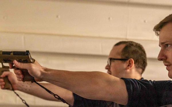 Two individuals in dark navy blue uniforms are engaged in what appears to be a firearms training exercise at an indoor shooting range. The person in the foreground is aiming a tan/FDE-colored semi-automatic pistol with a tethered cord attached, while a second uniformed individual observes from behind. Both wear name tags and insignia consistent with U.S. Navy personnel.