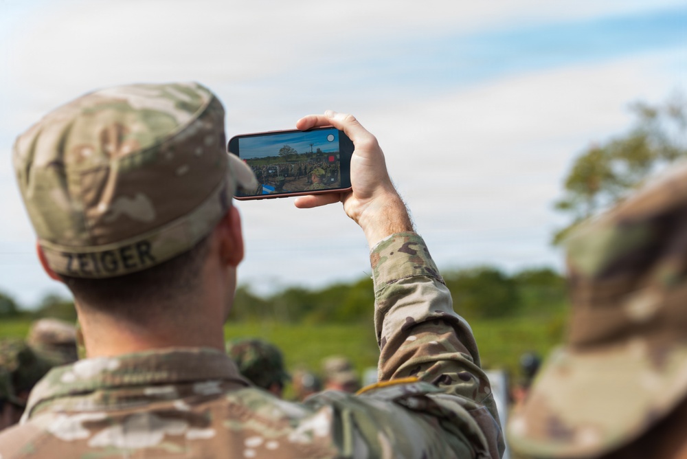 A soldier named Zeiger, wearing OCP (Operational Camouflage Pattern) uniform and patrol cap, is photographing or recording a military event or gathering with a smartphone held in landscape orientation. The phone screen shows a crowd of uniformed personnel in an outdoor field setting. A second soldier in similar uniform is partially visible in the foreground right.