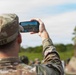 A soldier named Zeiger, wearing OCP (Operational Camouflage Pattern) uniform and patrol cap, is photographing or recording a military event or gathering with a smartphone held in landscape orientation. The phone screen shows a crowd of uniformed personnel in an outdoor field setting. A second soldier in similar uniform is partially visible in the foreground right.
