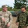 Two military personnel in different camouflage uniforms are engaged in conversation in an outdoor field setting. The soldier on the left is identified by his name tape as 'QUINONES' and wears a U.S. Army OCP (Operational Camouflage Pattern) uniform, while the soldier on the right wears what appears to be a Japan Ground Self-Defense Force (JGSDF) camouflage uniform. Military vehicles are visible in the background, suggesting a joint training exercise or bilateral military engagement.