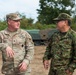 Two military personnel in different camouflage uniforms are engaged in conversation in an outdoor field setting. The soldier on the left is identified by his name tape as 'QUINONES' and wears a U.S. Army OCP (Operational Camouflage Pattern) uniform, while the soldier on the right wears what appears to be a Japan Ground Self-Defense Force (JGSDF) camouflage uniform. Military vehicles are visible in the background, suggesting a joint training exercise or bilateral military engagement.