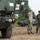 Two military personnel in different camouflage uniforms are standing and conversing near military vehicles on a dirt surface. One soldier wears a US Army MultiCam/OCP pattern uniform, while the other wears a distinct camouflage pattern consistent with the Japan Ground Self-Defense Force. The setting appears to be a joint military exercise or equipment demonstration, with tracked armored vehicles visible in the background.