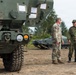 Two military personnel in different camouflage uniforms are standing and conversing near military vehicles on a dirt surface. One soldier wears a US Army MultiCam/OCP pattern uniform, while the other wears a distinct camouflage pattern consistent with the Japan Ground Self-Defense Force. The setting appears to be a joint military exercise or equipment demonstration, with tracked armored vehicles visible in the background.