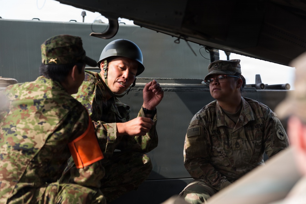 The image shows military personnel from at least two different armed forces engaged in what appears to be a briefing or discussion inside or alongside a military vehicle. One soldier wearing a black helmet and Japanese Ground Self-Defense Force (JGSDF) camouflage pattern uniform is speaking and gesturing, while a U.S. Army soldier in MultiCam/OCP uniform listens attentively. This appears to be a joint military exercise or training event between Japanese and American forces.