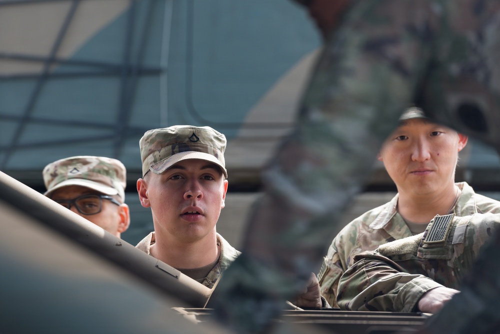 Three military personnel in U.S. Army Operational Camouflage Pattern (OCP) uniforms are visible, appearing to be positioned around or inside a military vehicle. The central figure wears a patrol cap with a rank insignia visible, while the soldier on the right displays a U.S. flag patch on the shoulder. The image is taken from a low angle through or around vehicle components, creating a framed perspective.