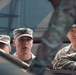 Three military personnel in U.S. Army Operational Camouflage Pattern (OCP) uniforms are visible, appearing to be positioned around or inside a military vehicle. The central figure wears a patrol cap with a rank insignia visible, while the soldier on the right displays a U.S. flag patch on the shoulder. The image is taken from a low angle through or around vehicle components, creating a framed perspective.