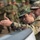 Two military personnel in camouflage uniforms are examining or discussing something in the foreground, likely a piece of equipment or a map surface. One soldier wears a distinctive green woodland-pattern cap with what appears to be a national emblem, while the other wears a MultiCam/OCP-pattern uniform with a shoulder patch. A group of soldiers in camouflage uniforms stands in the background, suggesting a training or briefing event.