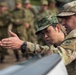 Two military personnel in camouflage uniforms are examining or discussing something in the foreground, likely a piece of equipment or a map surface. One soldier wears a distinctive green woodland-pattern cap with what appears to be a national emblem, while the other wears a MultiCam/OCP-pattern uniform with a shoulder patch. A group of soldiers in camouflage uniforms stands in the background, suggesting a training or briefing event.