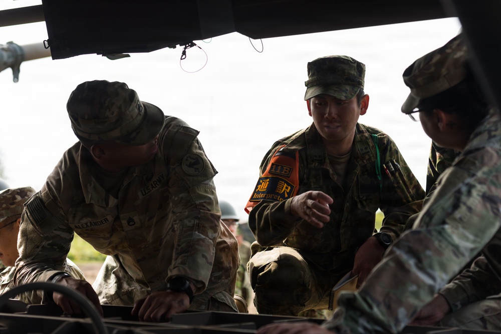 Three military personnel in camouflage uniforms are gathered around what appears to be a vehicle, reviewing documents or a map. The central figure is wearing an orange interpreter vest with Japanese characters (通訳/Interpreter) and a unit patch reading '11B', suggesting a joint US-Japanese military exercise. The soldier on the left has a visible name tape reading 'ALEXANDER' and 'US ARMY' patches.