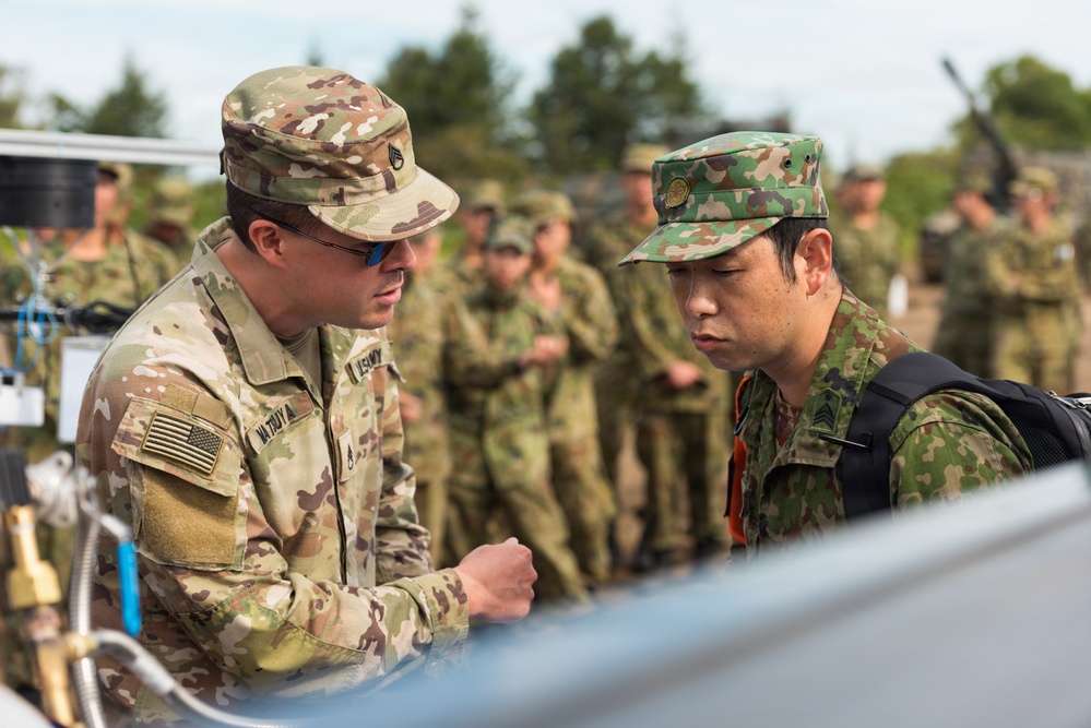 Two military personnel in different camouflage uniforms are closely examining equipment together in a field setting. The soldier on the left wears a US Army OCP (Operational Camouflage Pattern) uniform with an American flag patch, while the soldier on the right wears a Japanese Ground Self-Defense Force (JGSDF) camouflage uniform. A group of additional military personnel is visible in the background, suggesting a joint training exercise or demonstration.
