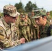 Two military personnel in different camouflage uniforms are closely examining equipment together in a field setting. The soldier on the left wears a US Army OCP (Operational Camouflage Pattern) uniform with an American flag patch, while the soldier on the right wears a Japanese Ground Self-Defense Force (JGSDF) camouflage uniform. A group of additional military personnel is visible in the background, suggesting a joint training exercise or demonstration.