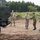 The image shows a military interaction between what appear to be U.S. Army soldiers in MultiCam/OCP uniforms and soldiers in a different camouflage pattern, likely Japanese Ground Self-Defense Force personnel, near a large military vehicle. Two groups are engaged in a handshake greeting, suggesting a joint exercise or bilateral military cooperation event. The setting is an outdoor training area or field site with dirt ground and forested background.
