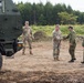 The image shows a military interaction between what appear to be U.S. Army soldiers in MultiCam/OCP uniforms and soldiers in a different camouflage pattern, likely Japanese Ground Self-Defense Force personnel, near a large military vehicle. Two groups are engaged in a handshake greeting, suggesting a joint exercise or bilateral military cooperation event. The setting is an outdoor training area or field site with dirt ground and forested background.