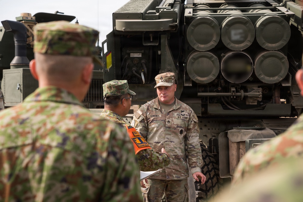 A U.S. Army soldier identified by nametape as 'QUINONES' appears to be conducting a briefing or demonstration in front of what appears to be an M270 Multiple Launch Rocket System (MLRS) or similar rocket artillery system. He is addressing a group that includes at least one soldier in a different camouflage pattern consistent with the Japan Ground Self-Defense Force uniform, suggesting a joint military exchange or training event. An orange armband worn by one of the foreign soldiers indicates an observer or liaison role.