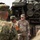 A U.S. Army soldier identified by nametape as 'QUINONES' appears to be conducting a briefing or demonstration in front of what appears to be an M270 Multiple Launch Rocket System (MLRS) or similar rocket artillery system. He is addressing a group that includes at least one soldier in a different camouflage pattern consistent with the Japan Ground Self-Defense Force uniform, suggesting a joint military exchange or training event. An orange armband worn by one of the foreign soldiers indicates an observer or liaison role.