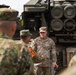 A U.S. Army soldier identified by nametape as 'QUINONES' appears to be conducting a briefing or demonstration in front of what appears to be an M270 Multiple Launch Rocket System (MLRS) or similar rocket artillery system. He is addressing a group that includes at least one soldier in a different camouflage pattern consistent with the Japan Ground Self-Defense Force uniform, suggesting a joint military exchange or training event. An orange armband worn by one of the foreign soldiers indicates an observer or liaison role.