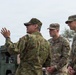 Three military personnel in camouflage uniforms are engaged in a discussion outdoors near military vehicles. One individual, wearing a distinctive green camouflage pattern different from the other two, is gesturing with his hand while speaking to two U.S. Army soldiers. The setting appears to be a military exercise or joint operations event.
