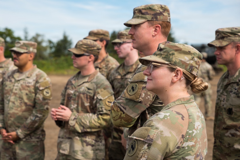 A group of U.S. Army soldiers in Operational Camouflage Pattern (OCP) uniforms are standing in formation outdoors, appearing to observe or listen to a briefing or ceremony. The soldiers wear patrol caps and some have protective eyewear. The setting appears to be a training area or field environment with trees and overcast sky visible in the background.