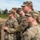 A group of U.S. Army soldiers in Operational Camouflage Pattern (OCP) uniforms are standing in formation outdoors, appearing to observe or listen to a briefing or ceremony. The soldiers wear patrol caps and some have protective eyewear. The setting appears to be a training area or field environment with trees and overcast sky visible in the background.