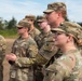 A group of U.S. Army soldiers in Operational Camouflage Pattern (OCP) uniforms are standing in formation outdoors, appearing to observe or listen to a briefing or ceremony. The soldiers wear patrol caps and some have protective eyewear. The setting appears to be a training area or field environment with trees and overcast sky visible in the background.