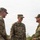 Three military personnel in camouflage uniforms are engaged in a friendly conversation outdoors. Two individuals wear U.S. Army OCP (Operational Camouflage Pattern) uniforms, while the third wears what appears to be a Japanese Ground Self-Defense Force uniform, suggesting a multinational military interaction. The atmosphere appears relaxed and collegial.