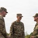 Three military personnel in camouflage uniforms are engaged in a friendly conversation outdoors. Two individuals wear U.S. Army OCP (Operational Camouflage Pattern) uniforms, while the third wears what appears to be a Japanese Ground Self-Defense Force uniform, suggesting a multinational military interaction. The atmosphere appears relaxed and collegial.